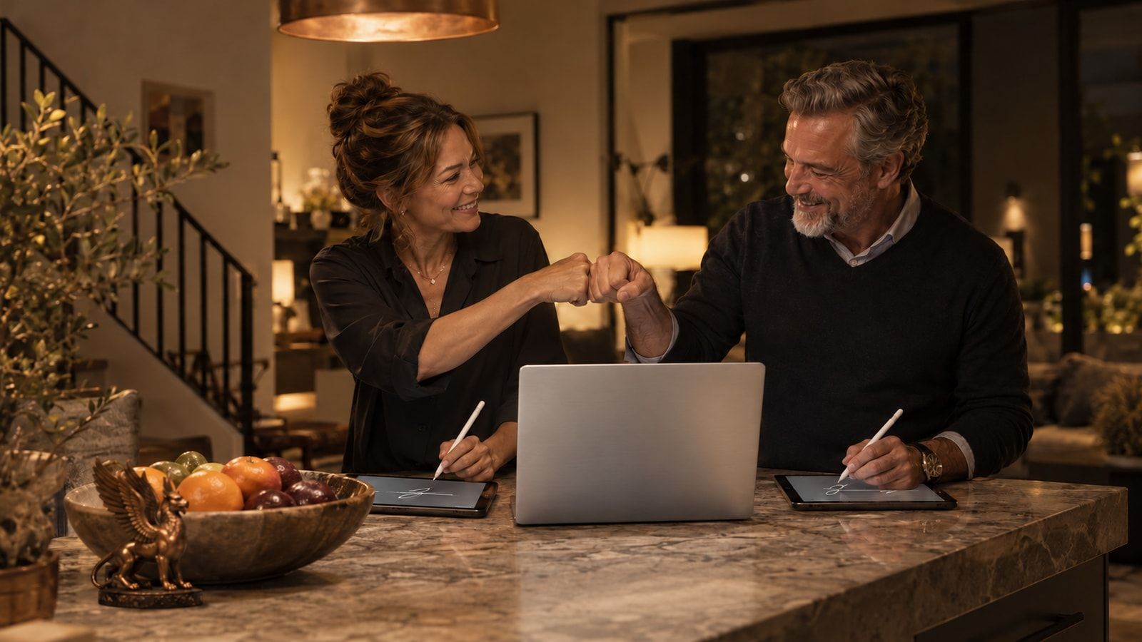Two adults fist-bumping while holding styluses over tablets at a kitchen island.
